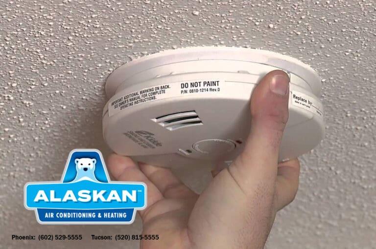 hand holding fire detector near ceiling Alaskan Air Conditioning and Heating logo and contact phone numbers