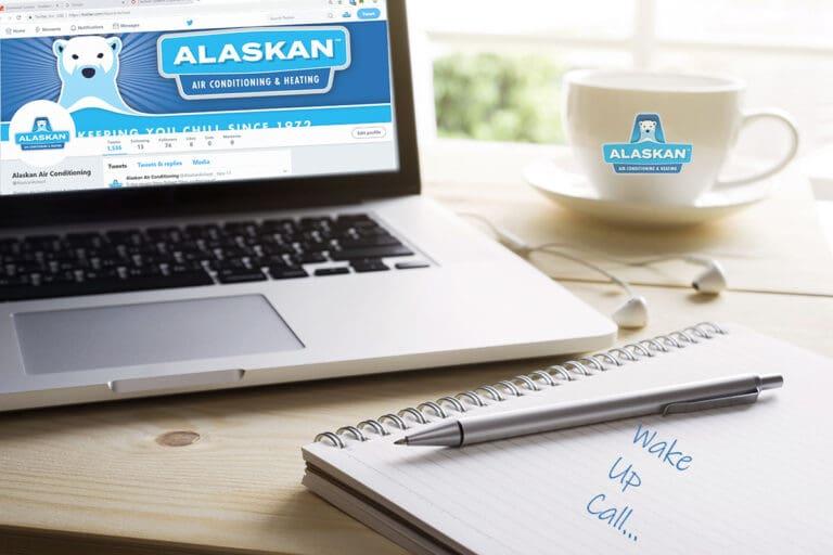 Desk view of laptop showing Alaskan website, coffee cup with Alaskan logo, and note book and pen with note "wake up call..."