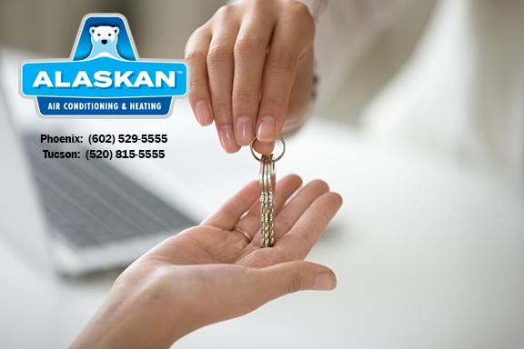 One hand giving another open hand a set of keys, laptop open in background, with Alaskan Air Conditioning and Heating logo and contact phone numbers in top left corner