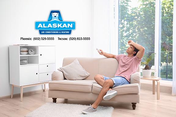 man sitting on couch, hand to forehead, holding remote, Alaskan Air Conditioning and Heating logo and contact phone numbers in top left quadrant