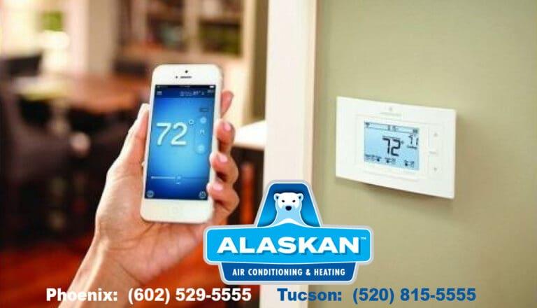 wall mounted thermostat with hand holding phone with matching thermostat app open with Alaskan Air Conditioning and Heating logo and contact phone numbers across bottom